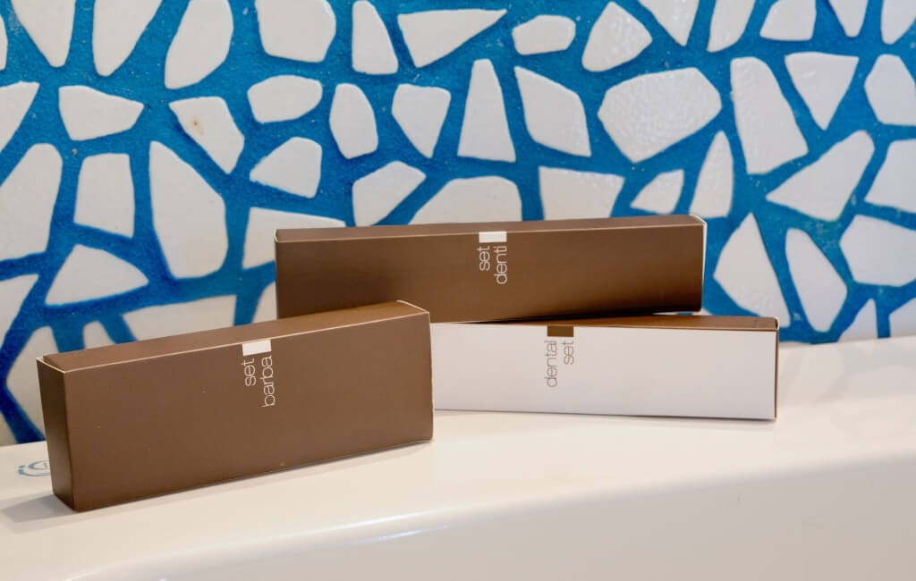 Drei braune und weiße Boxen auf einer Badewanne, beschriftet mit "set barba", "set denti" und "dental set", vor einer blauen Wand.