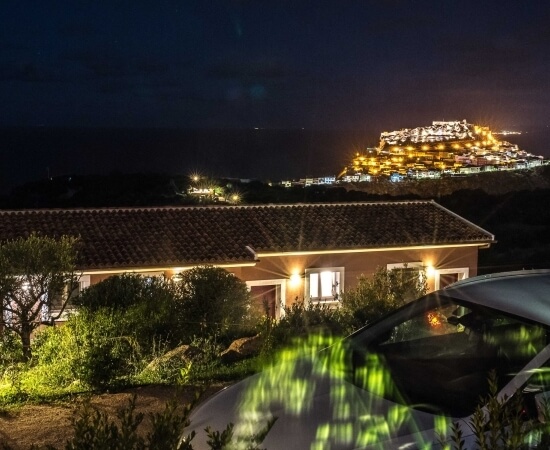 A radiant white house at night, surrounded by shrubs, with glowing windows and a distant illuminated house on a hillside.