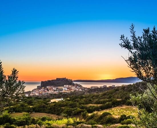 Serene landscape of a hilltop town with white architecture, lush fields, water, and a mountain at sunset.