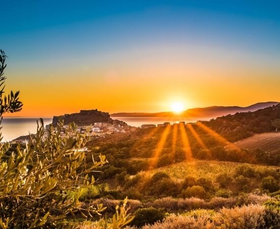 Serene sunset over water with hills, a village, and trees, featuring a warm glow from the sun's rays.
