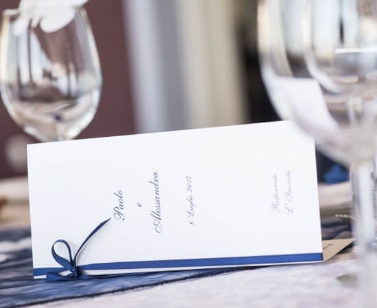 Invito di nozze elegante con nastro blu, posizionato su un tavolo con bicchieri di vino sullo sfondo sfocato.
