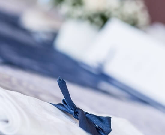Nastro blu legato a un fiocco su una tovaglia bianca, con sfondo sfocato di fiori, suggerendo un'atmosfera elegante.