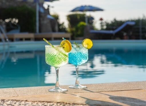 Deux cocktails colorés sur le bord d'une piscine, garnis de fruits et d'herbes, entourés de chaises longues et de buissons.