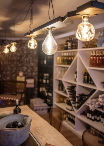 Bar accogliente con bancone in legno chiaro, tubi metallici con bottiglie scure e luci sospese che creano un'atmosfera intima.