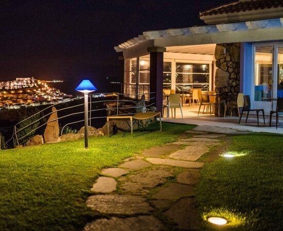 A serene nighttime scene of a house with a patio overlooking the ocean and a lit-up town in the background.
