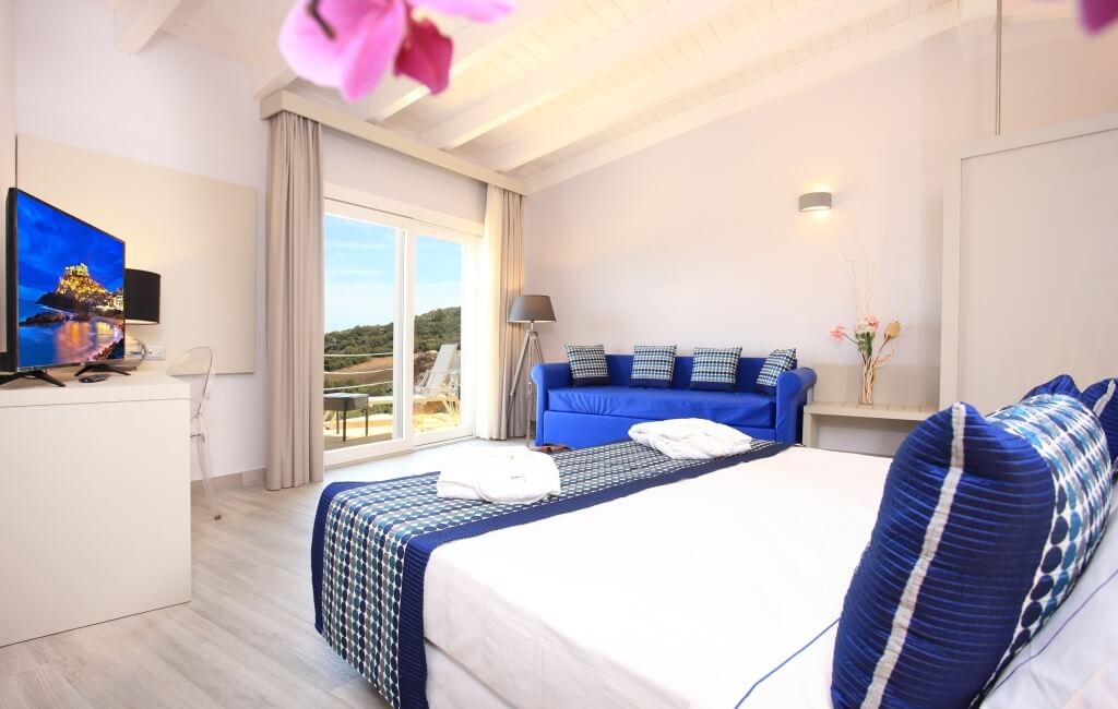 Ein einladendes Hotelzimmer mit einem Doppelbett, blauen Kissen, Sofa, Fensterblick und warmem Holzfußboden.