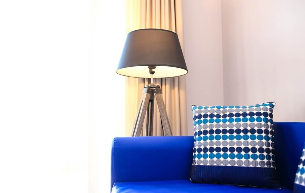 Blaues Sofa mit gepunktetem Kissen, Lampe und beiger Gardine vor einem Fenster in einem einladenden Wohnzimmer.