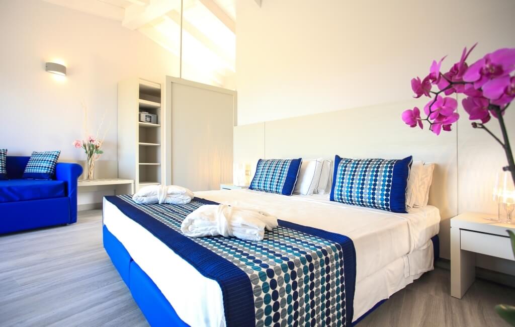 Ein einladendes Hotelzimmer in Weiß mit blauen und rosa Akzenten, gemütlichem Bett, Couch und Blumenarrangements.