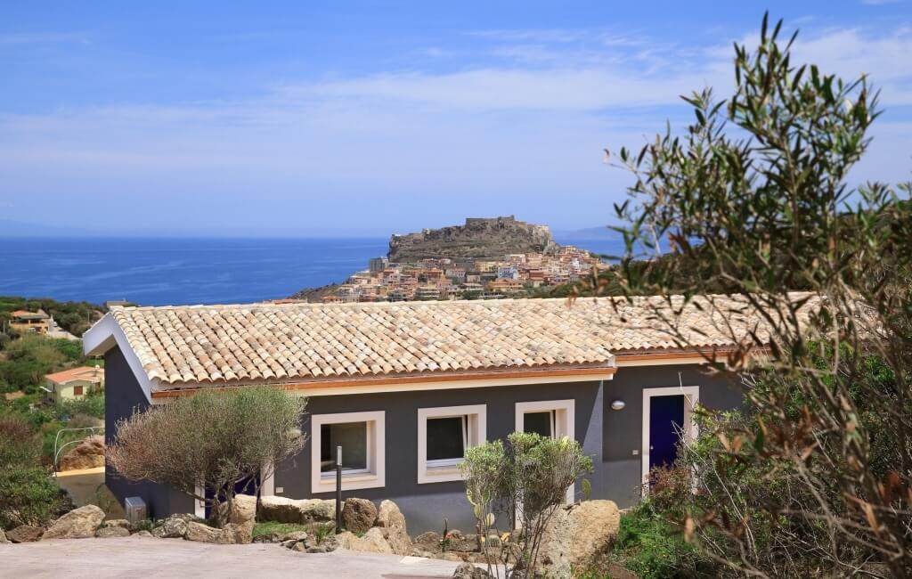 Ein graues Haus mit weißem Trim, blauer Tür, vor Wasser und einer Insel, umgeben von Bäumen und Felsen, unter blauem Himmel.