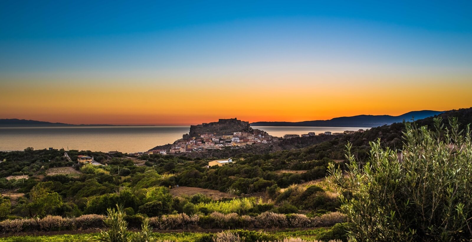 Serene coastal scene with a hillside town, white and brown buildings, lush greenery, and a colorful sunrise/sunset sky.