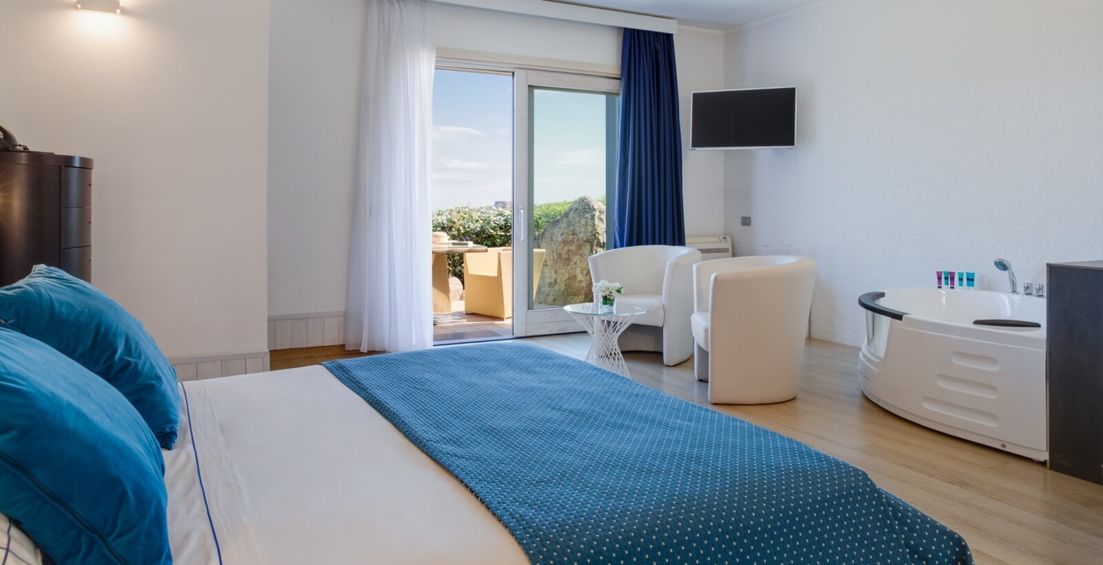 Moderne Hotelzimmer mit großem Bett, weißen Stühlen, Flachbild-TV, Badewanne und viel natürlichem Licht.