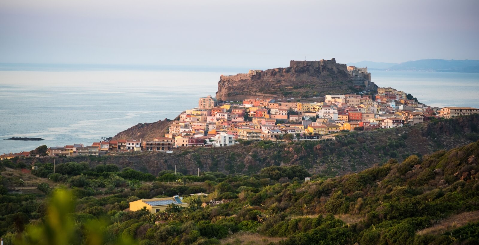 Malerische Küstenstadt auf einem Hügel, mit Häusern, Bäumen, einem Solarpanel und einem großen Gewässer im Hintergrund.