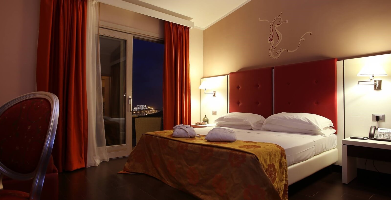 Hotelzimmer mit Bett, Nachttisch und rotem Stuhl, dekoriert in Rot und Weiß, mit Fenster und nächtlicher Stadtansicht.