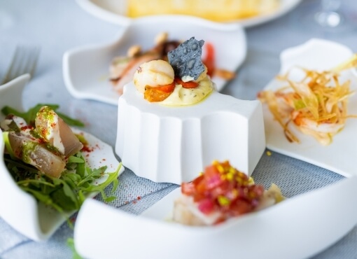 Collection de petits plats blancs sur une nappe bleu clair, contenant divers aliments, dont une salade et une sauce.