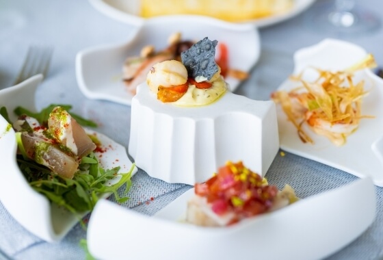 Una selezione di piccoli piatti bianchi con cibo elegante su una tovaglia blu, suggerendo un ambiente di ristorazione formale.