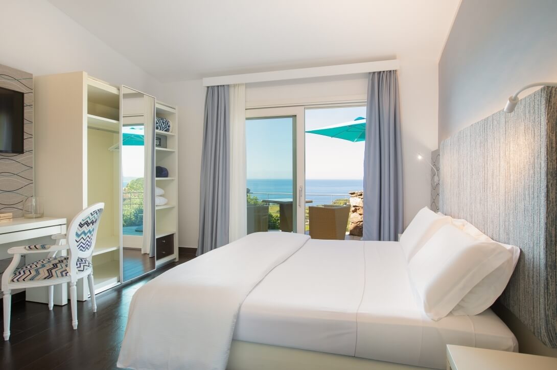 Camera d'hotel serena con vista sull'oceano, letto matrimoniale, scrivania e balcone con tavolo e sedie sotto un ombrello verde.