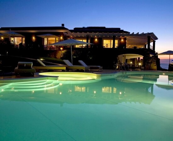 Large hillside house with a pool and patio, illuminated at dusk under a clear sky, creating a serene and inviting atmosphere.