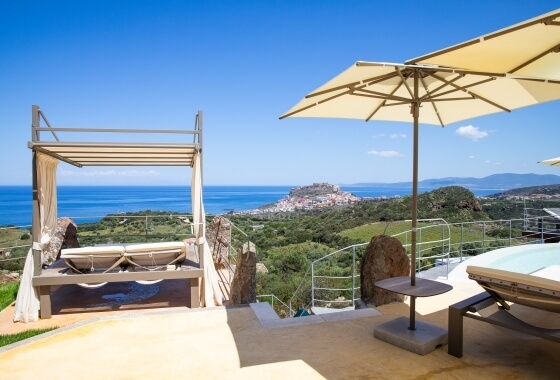 Panorama marino con patio, chaise lounge, ombrelloni, collina verde e cielo blu con nuvole sparse.