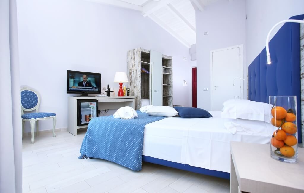 Schlafzimmer mit blau-weißer Farbgebung, großem Bett, Schreibtisch, Fernseher, Nachttisch und weißen Fliesenboden.