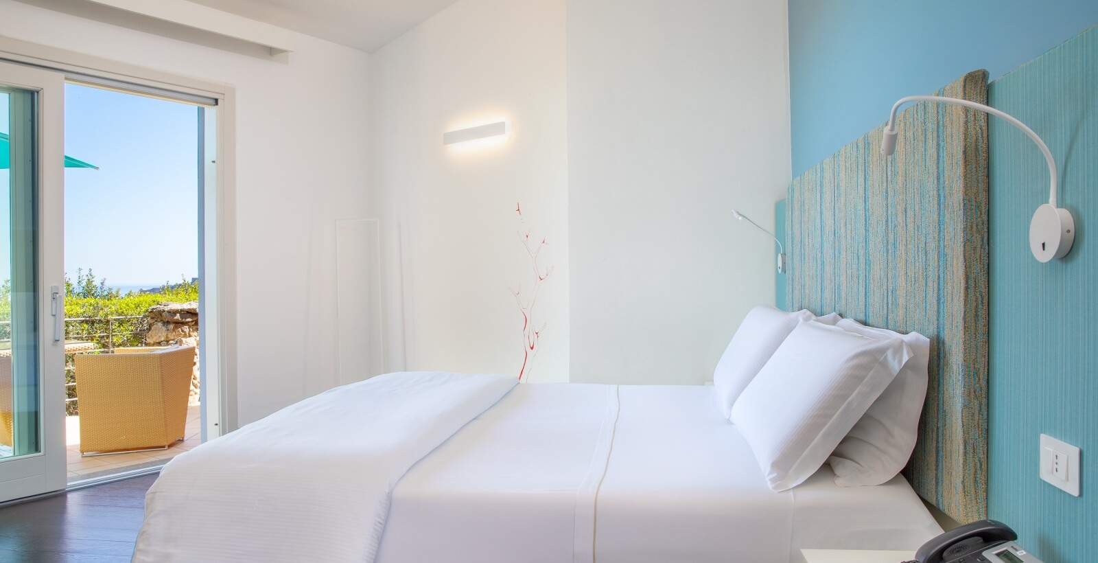 Camera moderna e serena con letto bianco, balcone, e dettagli blu e verde, che invita al relax.
