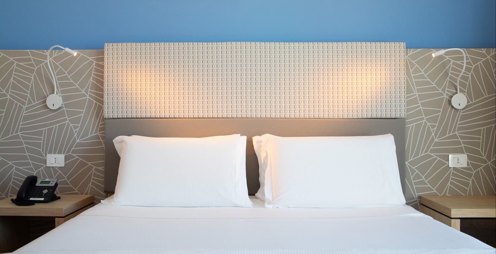 Camera da letto con letto bianco, comodini in legno, lampade regolabili e testiera decorativa su parete blu.