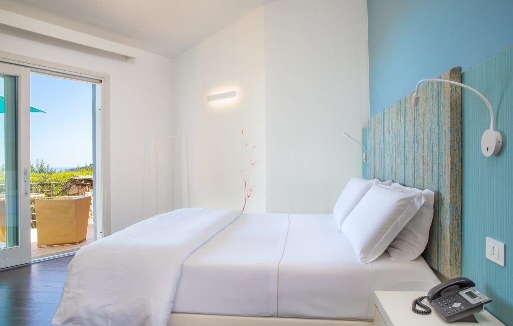 Ein ruhiges Schlafzimmer mit einem Bett, Nachttisch und einer Schiebetür zur Terrasse mit Blick auf üppiges Grün.