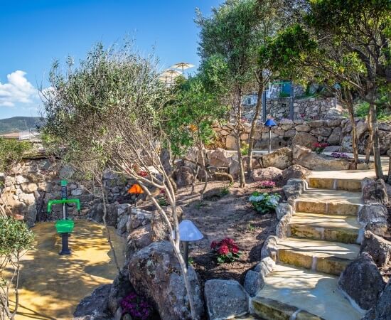 Serene park scene with stone staircase, vibrant flowers, yellow-tiled area, and green fountain, surrounded by trees and a stone wall.