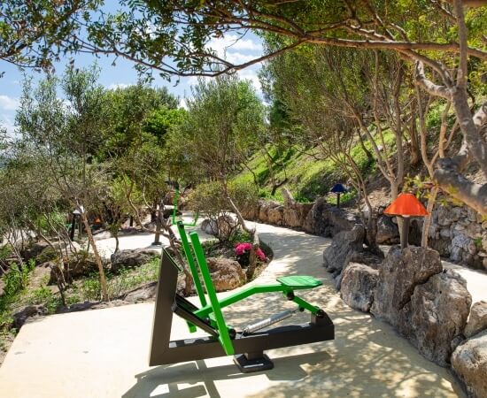 Serene outdoor scene with a concrete path, trees, rocks, and a green exercise machine under a blue sky with clouds.