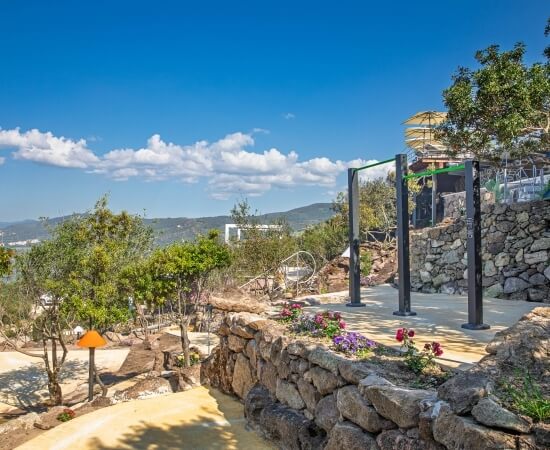 Scena serena all'aperto con un muro di pietra, fiori, struttura metallica, acqua sullo sfondo e montagne lontane.