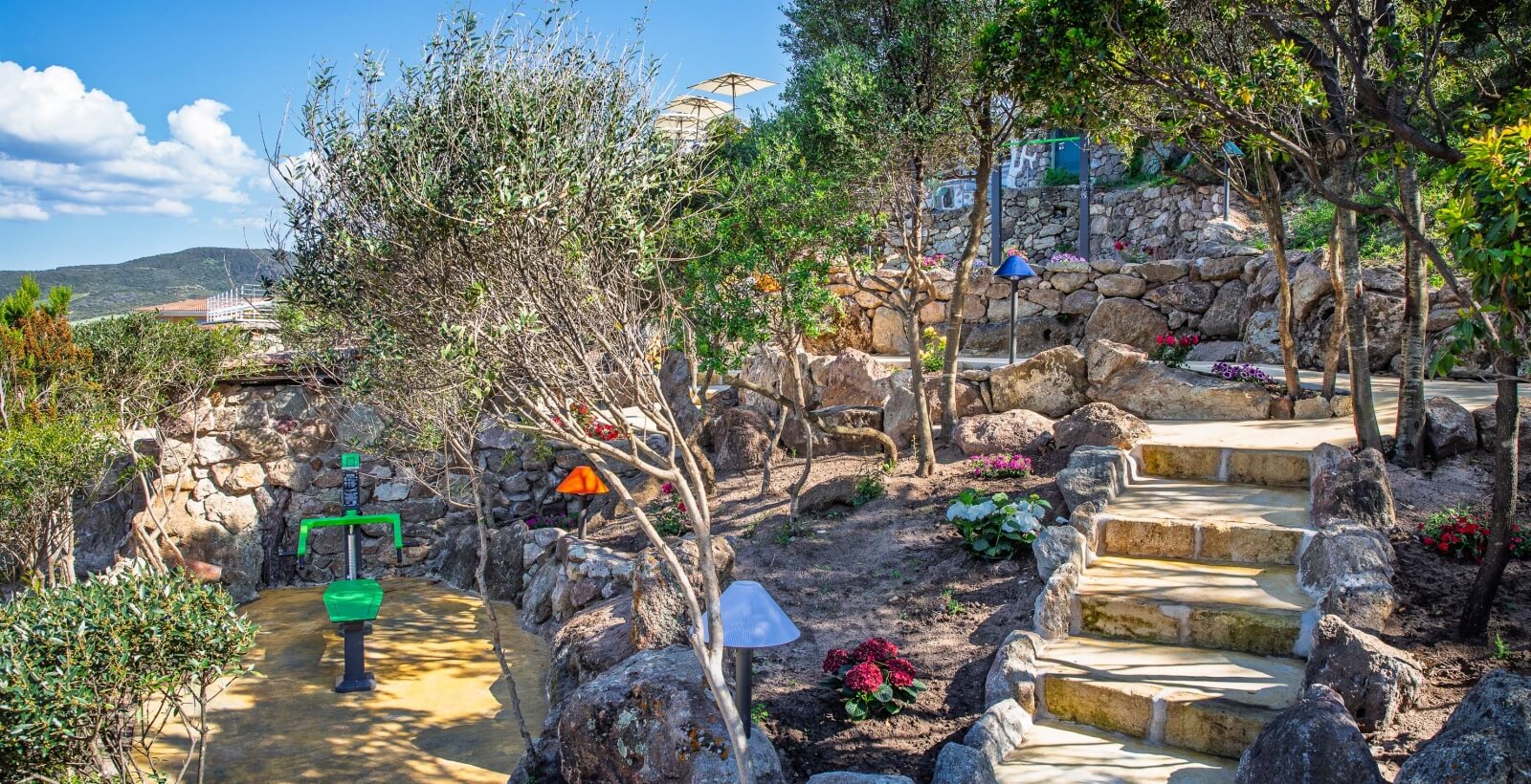 Serene outdoor scene with a stone staircase, walkway, lush greenery, a stream, and distant structures under a blue sky.