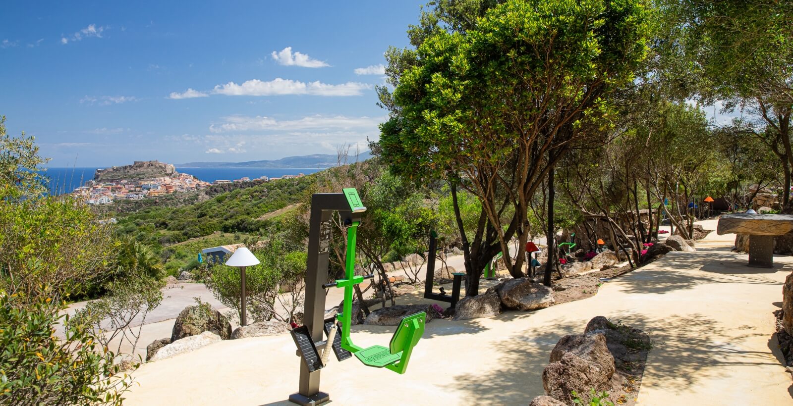 Area fitness all'aperto con macchina verde, alberi, attrezzature, rocce e vista sulla costa sotto un cielo soleggiato.