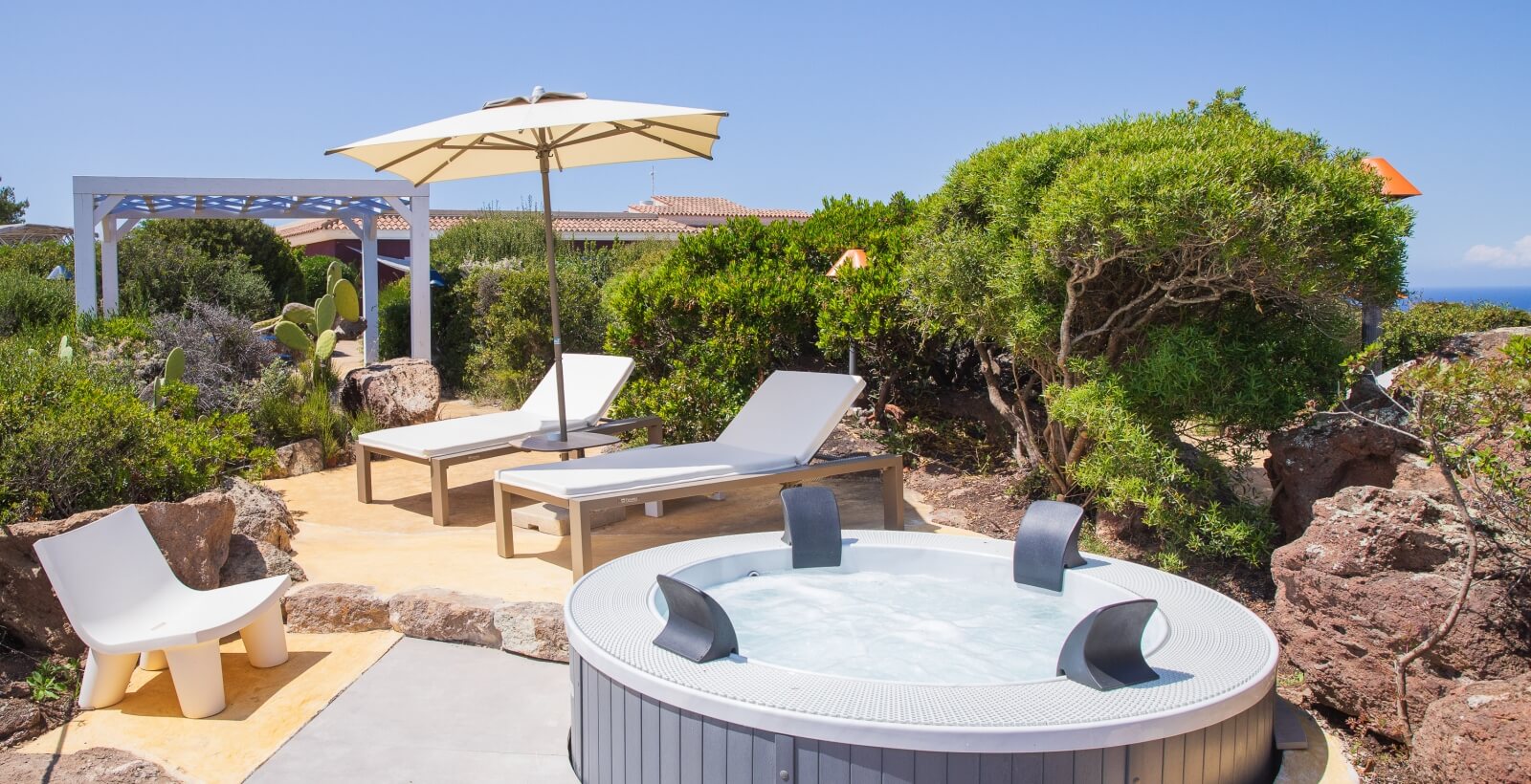 Impostazione serena all'aperto con vasca idromassaggio, lettini, ombrellone, rocce e pergola sullo sfondo.