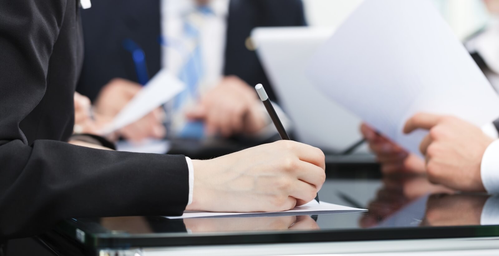 A person in a black suit writes on paper at a glass table, with blurred colleagues engaged in various tasks in the background.