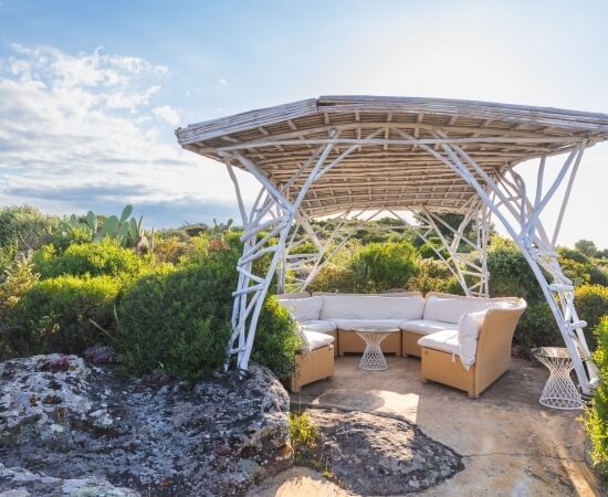 Area esterna serena con gazebo bianco, divano chiaro e piante, sotto un cielo blu punteggiato di nuvole bianche.
