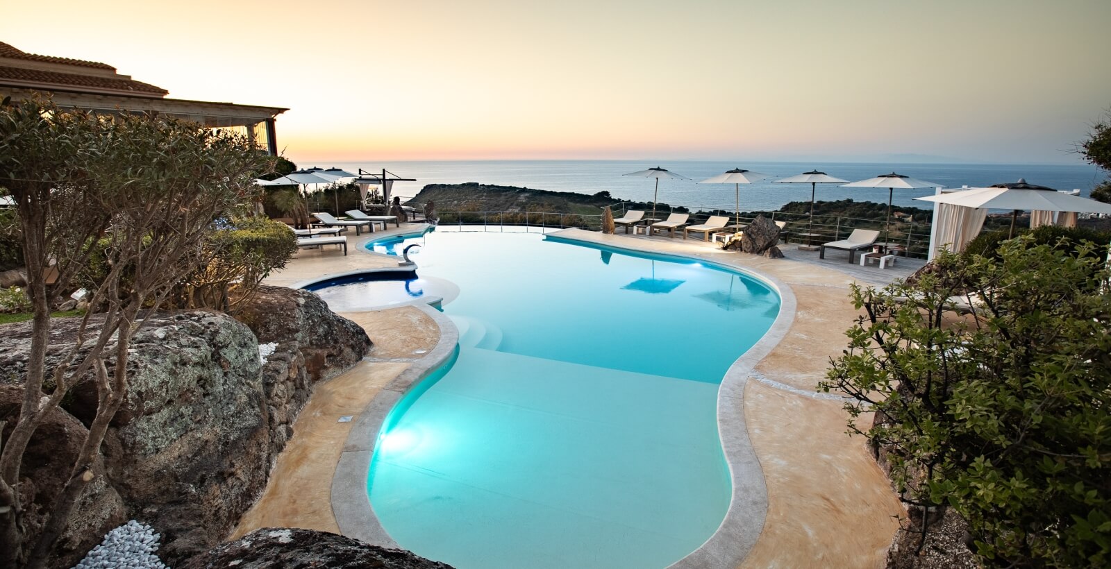 Ein ruhiger Poolbereich bei Sonnenuntergang mit einem großen, geschwungenen Pool, Liegen und umgebenden Bäumen.