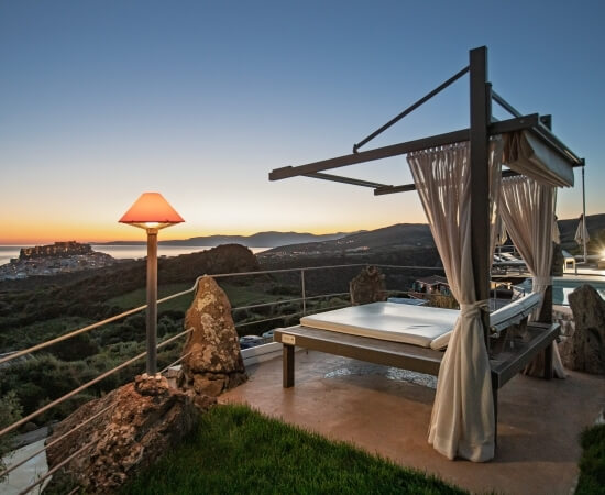 Ein Tagesbett mit Vorhängen und einer Lampe auf einer erhöhten Terrasse mit Blick auf eine atemberaubende Landschaft bei Sonnenuntergang.