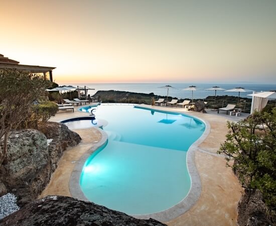 Ein ruhiger Pool bei Sonnenuntergang, umgeben von Felsen, Grün und Blick auf das Meer, mit Liegen und einem Cabana.
