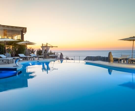 Ein ruhiger Poolbereich bei Sonnenuntergang, mit einem großen Pool, Liegen, einem Gebäude und dem Ozean im Hintergrund.