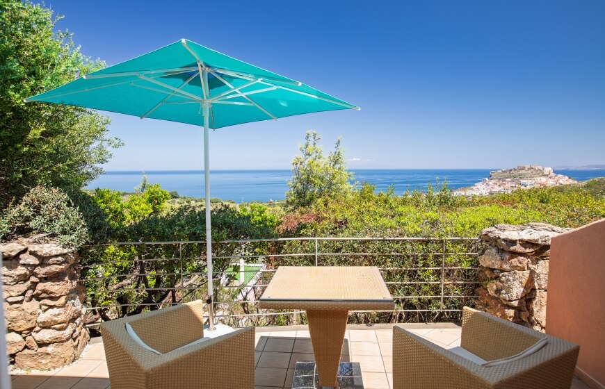 Patio con tavolo e sedie in rattan, ombreggiato da un ombrello, affacciato su un corpo d'acqua tranquillo e verdeggianti.