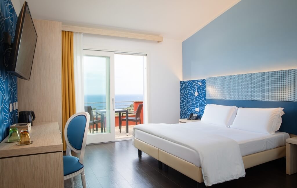 Modernes Hotelzimmer mit Bett, Schreibtisch, Fernseher und Balkon, dekoriert in Weiß und Blau, sauber und gut gepflegt.