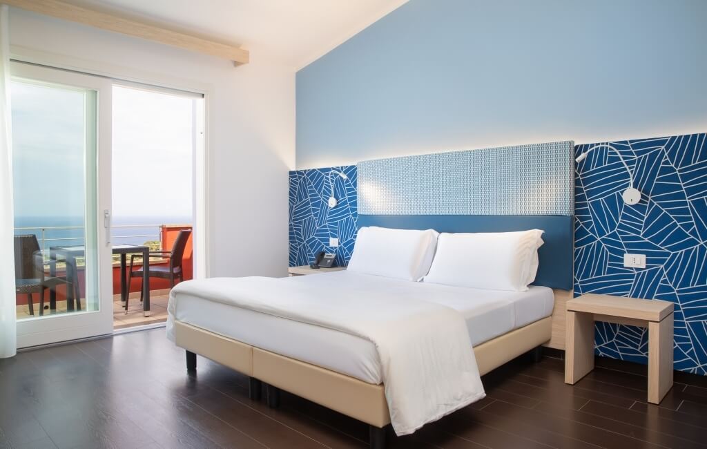 Ein einladendes Hotelzimmer mit einem Bett, Nachttisch, offener Glastür zum Balkon und warmem Holzboden.