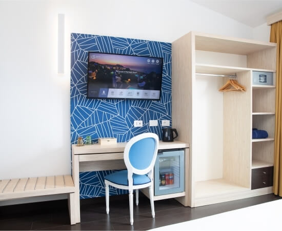 Hotelzimmer mit Schreibtisch, Stuhl, Fernseher, offener Kleiderschrank und dunklem Holzboden. Wand in Blau mit weißen Linien.