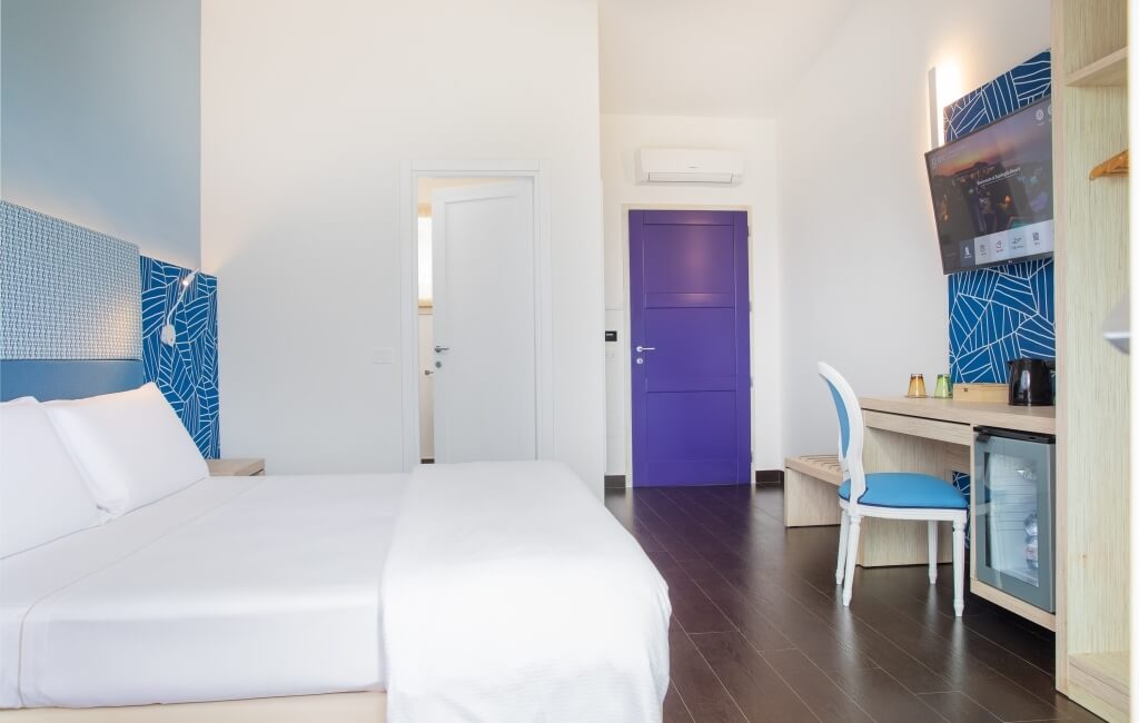 Ein modernes Hotelzimmer mit einem Bett, Schreibtisch, Fernseher und einer lila Tür, dekoriert in Weiß und Blau.