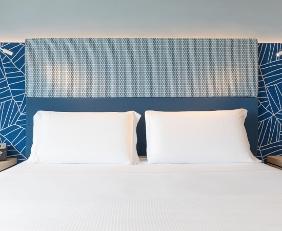 Doppelbett mit blau-weißer Kopfteil, weißen Kissen, blauen Wanddekorationen und grauer Wand im Hintergrund.