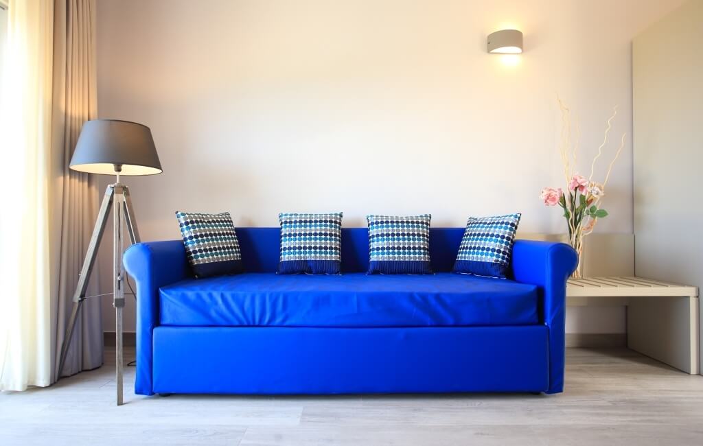 Ein Raum mit einem blauen Sofa, einer Stehlampe, einem Tisch und einer großen Fenster mit beigen Vorhängen.