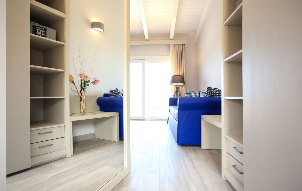 Heller Raum mit weißem Farbkonzept, Kleiderschrank links, blaue Couch rechts, Vase mit rosa Blumen und Fenster mit Vorhang.