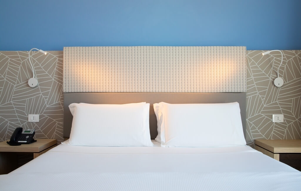 Schlafzimmer mit Bett, weißen Bettwaren, Nachttischen aus hellem Holz und blauer sowie grauer Wandgestaltung.