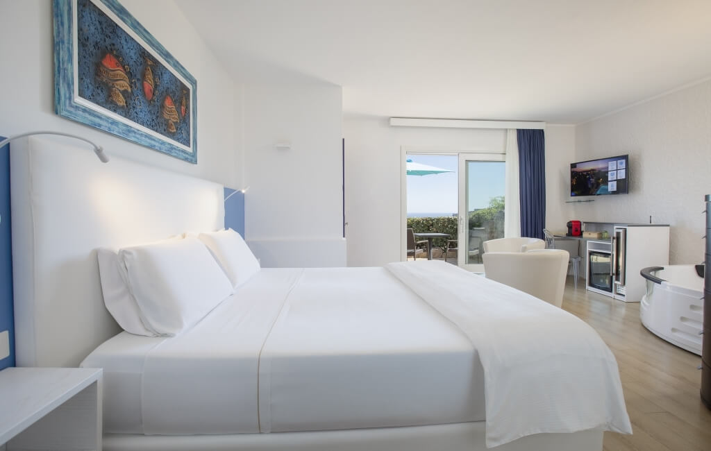 Ein modernes Hotelzimmer mit einem weißen Bett, Nachttisch, TV, und einer Glastür, die zu einer Terrasse führt.