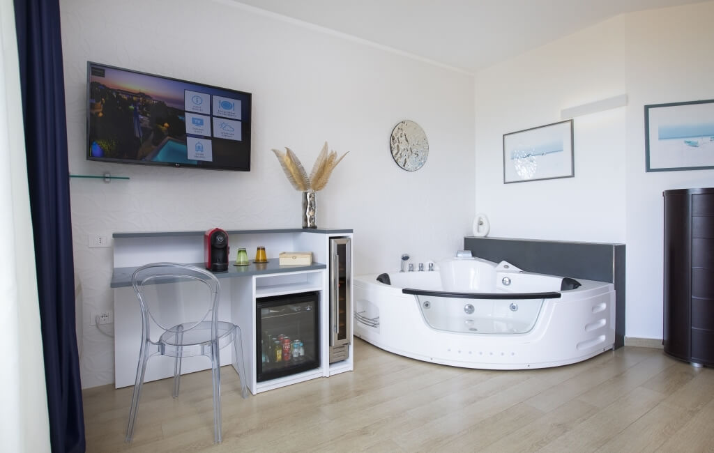 Modernes, weißes Hotelzimmer mit rundem Whirlpool, TV, Schreibtisch, Kunstwerken und einem warmen Holzschrank.