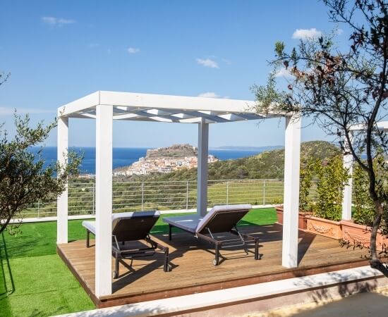Ein weißer Pergola mit Holzdeck und zwei Liegestühlen, umgeben von grünen Hügeln und einem Gewässer unter blauem Himmel.